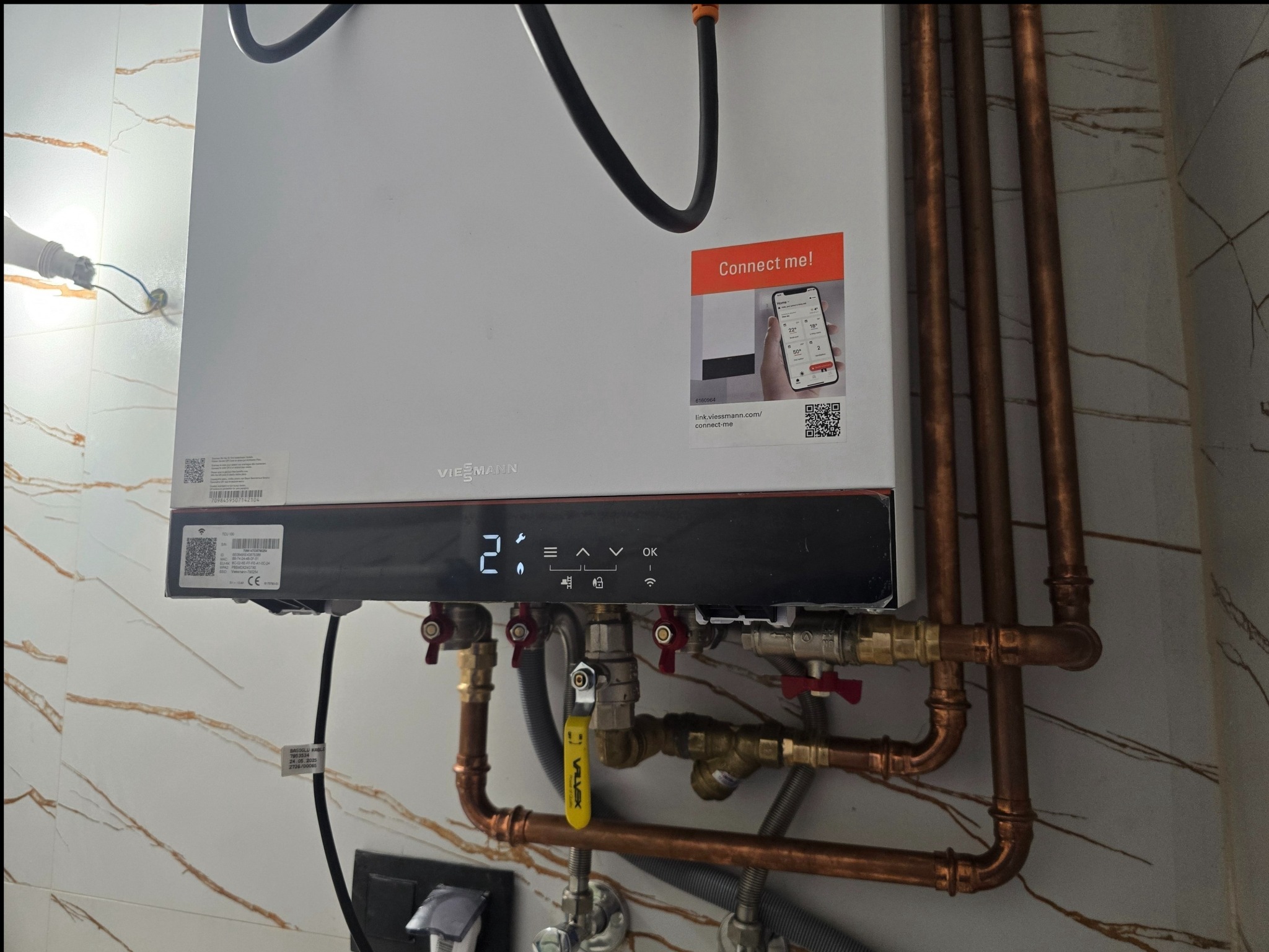 Nowoczesny, biały piec gazowy Viessmann z miedzianymi rurami instalacji, zamontowany na ścianie z marmurowym wzorem. Widoczny wyświetlacz i naklejka 'Connect me!'.