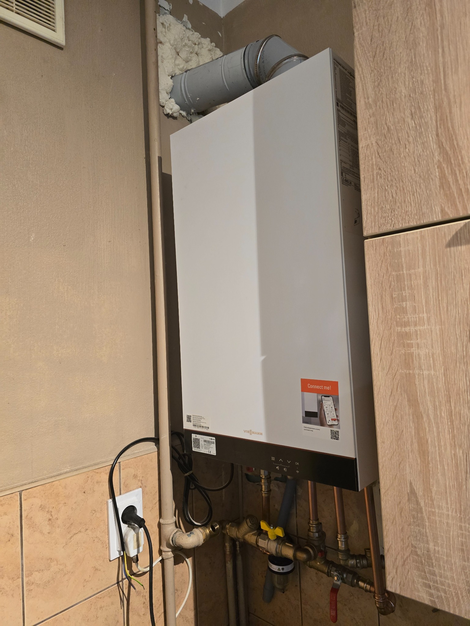 Biały piec gazowy Viessmann zamontowany na ścianie z widocznymi rurami miedzianymi, zaworami i podłączeniem elektrycznym. Ściana wykończona płytkami i panelami.