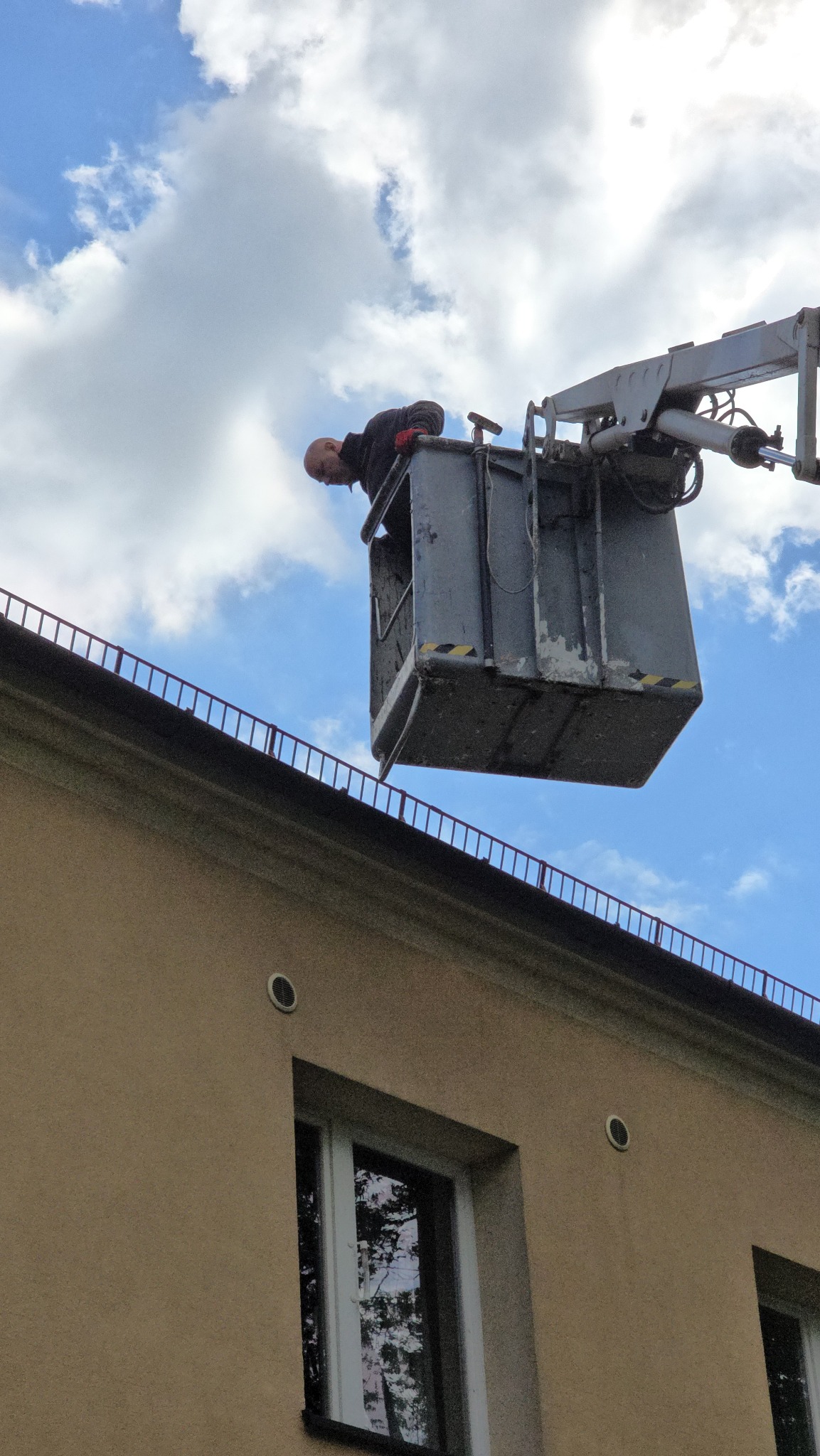 Pracownik w koszu podnośnika kontroluje dach budynku. Widoczne okna i błękitne niebo z chmurami. Nietypowy kadr z dołu.