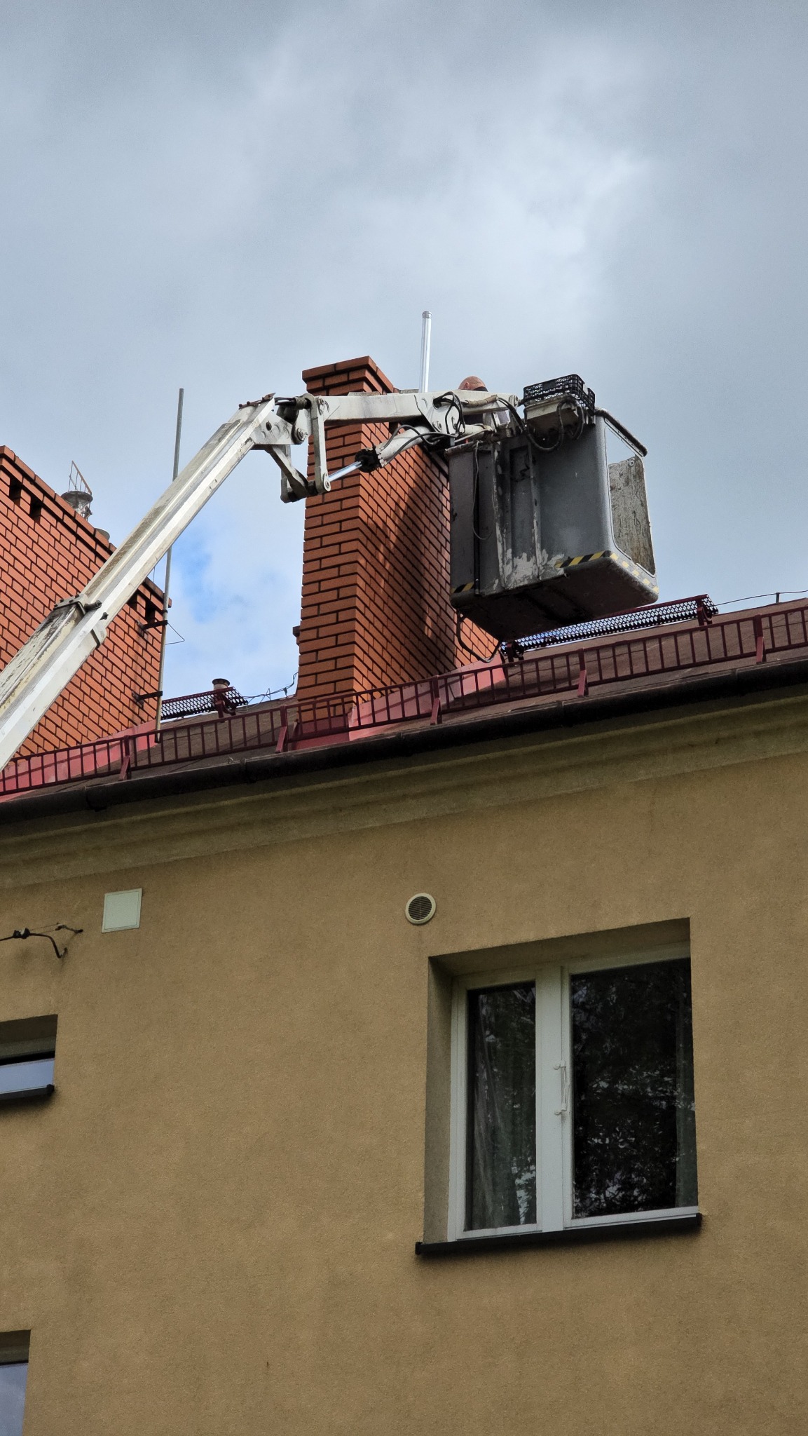 Podnośnik koszowy przy kominie na dachu budynku. Prace na wysokościach, widoczny fragment elewacji i okno. Szaro-białe niebo w tle.