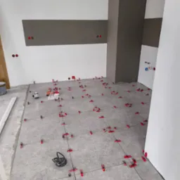 Szare płytki podłogowe w trakcie układania, z użyciem czerwonych klipsów poziomujących i lasera krzyżowego, widoczne puszki elektryczne na ścianie.