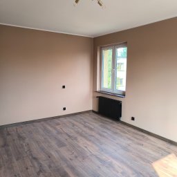 OLEKBUD OLEKSANDR REVIAKIN - Pusty pokój po odświeżeniu: ściany pomalowane na ciepły beż, ciemne panele podłogowe, białe okno z czarnym grzejnikiem pod spodem, złoty żyrandol na suficie, widoczne gniazdka elektryczne.