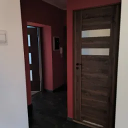 Korytarz po remoncie z ciemną podłogą z płytek i dwiema parami drzwi, z kt&oacute;rych jedne są drewniane z trzema poziomymi przeszkleniami.