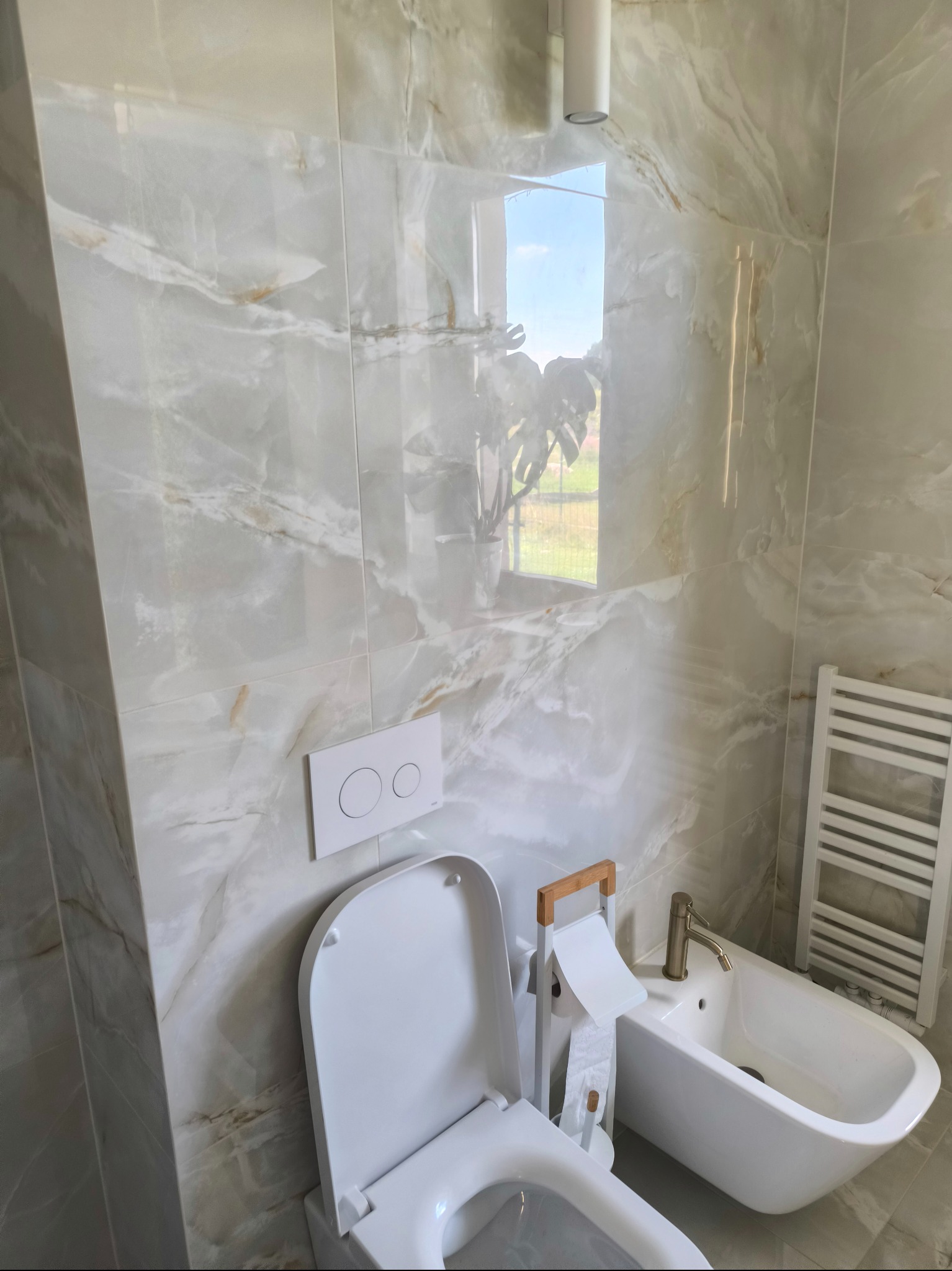 Wykończona łazienka z marmurowymi płytkami, wiszącą toaletą z podwójnym przyciskiem, bidetem z baterią w kolorze szczotkowanego złota i grzejnikiem drabinkowym.