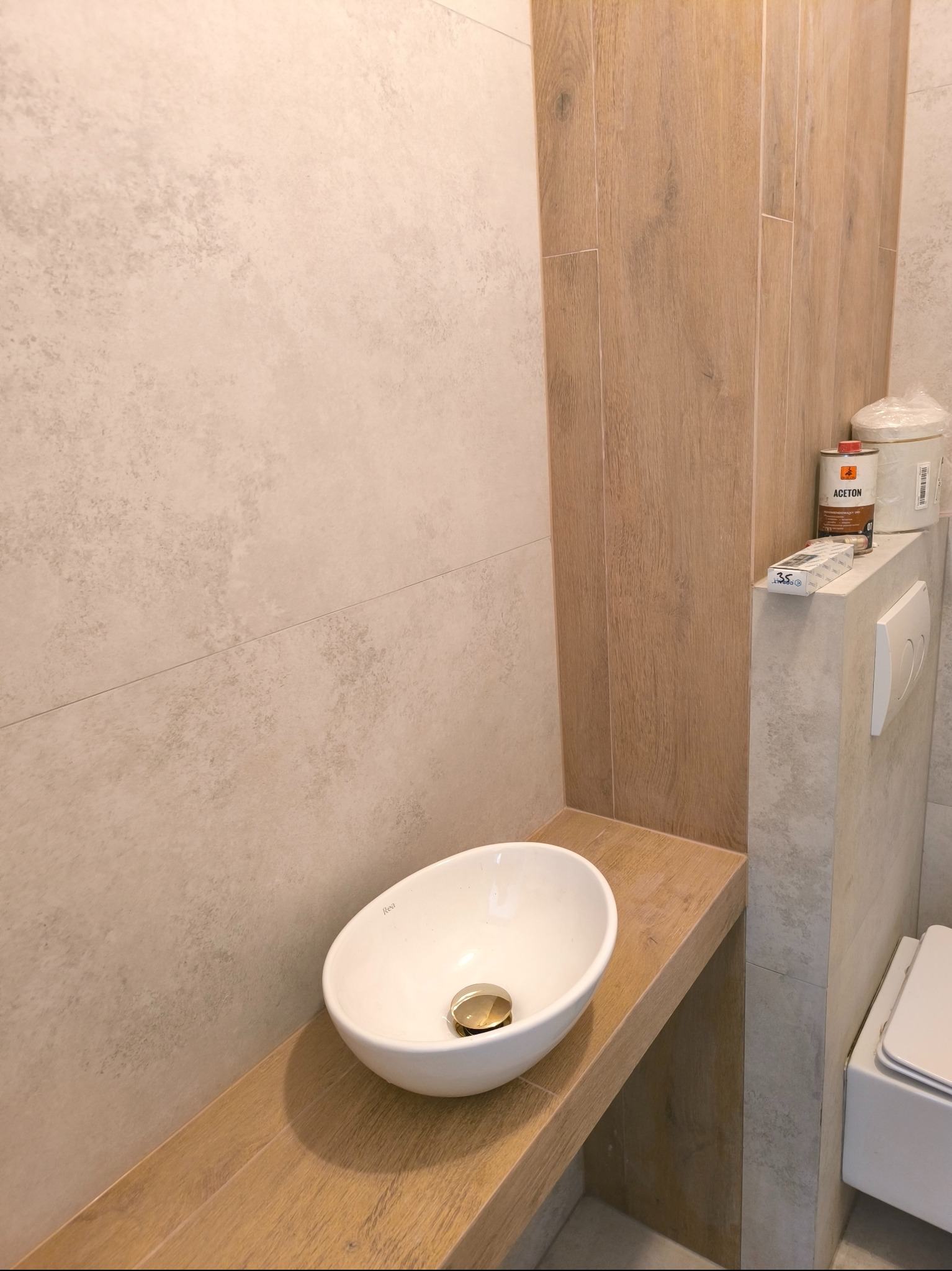 Nowoczesna łazienka w trakcie wykańczania: biała umywalka nablatowa na drewnopodobnym blacie, obok toaleta i płytki imitujące drewno, widoczne materiały budowlane i narzędzia.