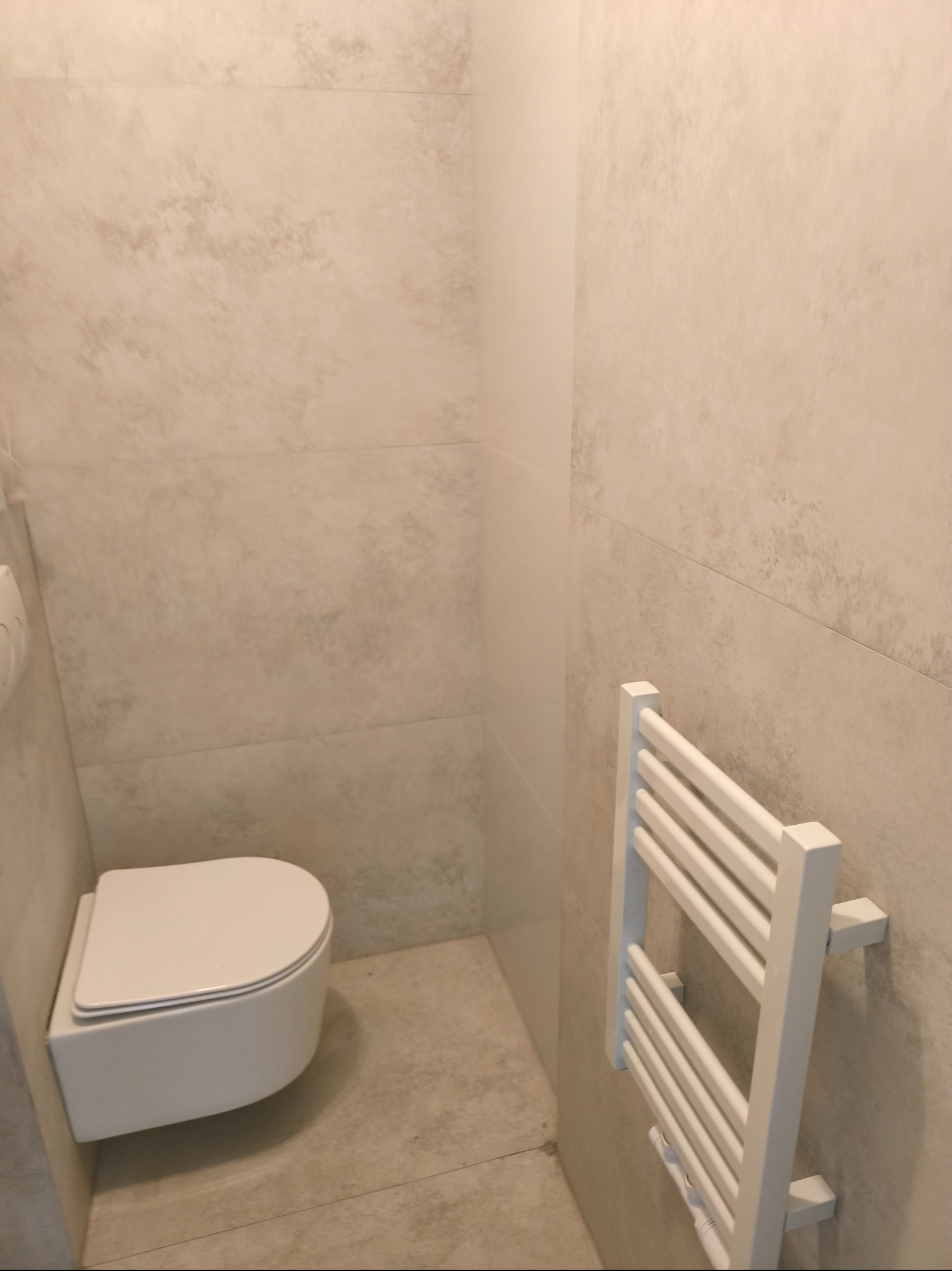 Nowoczesna łazienka z podwieszaną miską WC i białym grzejnikiem drabinkowym na ścianie z beżowymi płytkami imitującymi kamień.