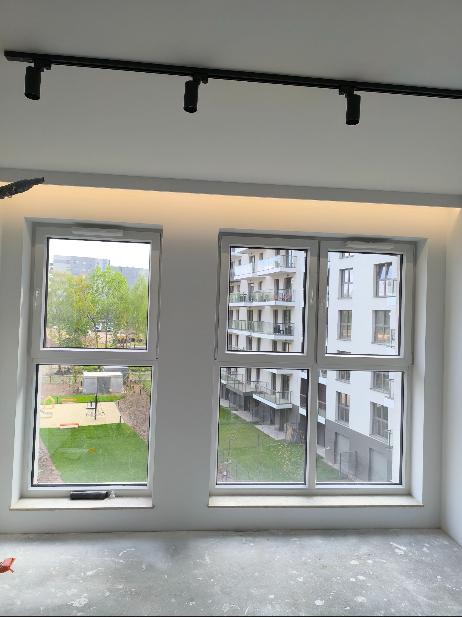 Wnętrze pokoju w trakcie prac wykończeniowych, widoczne dwa okna z widokiem na zewnątrz, surowa podłoga z plamami farby, oświetlenie szynowe na suficie.