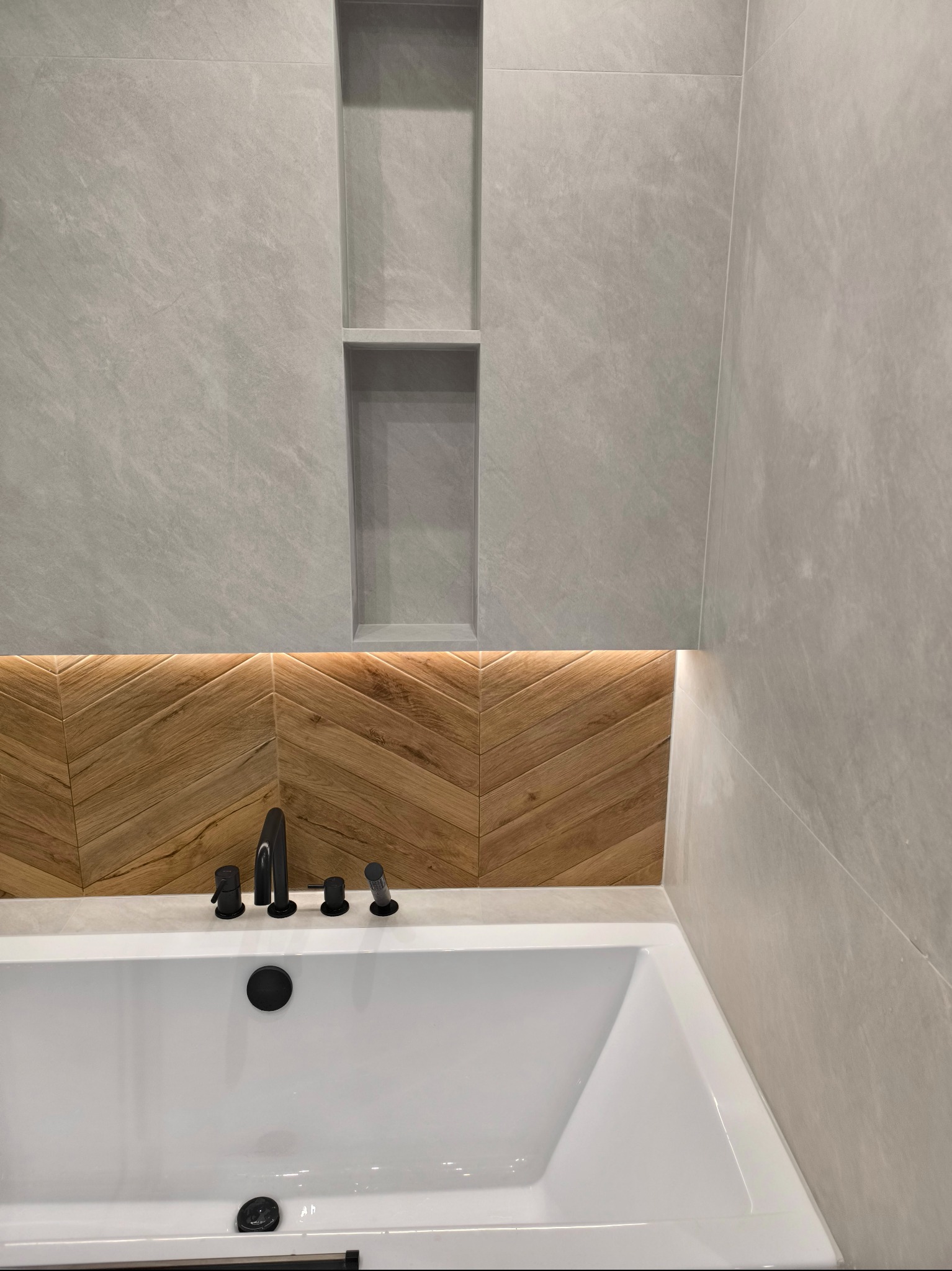 Nowoczesna łazienka z białą wanną, czarną armaturą, szarą płytką imitującą kamień i drewnianą mozaiką ułożoną w jodełkę z podświetleniem LED.