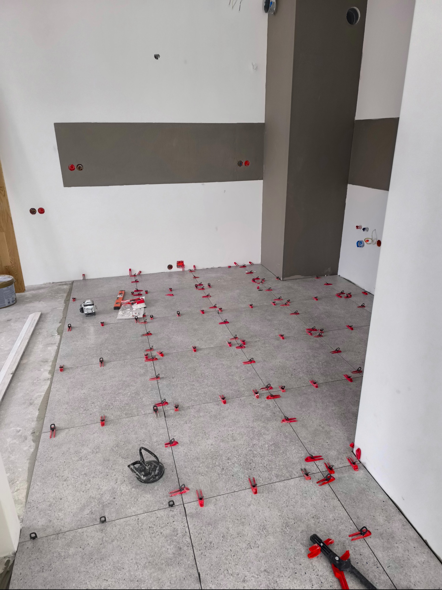 Szare płytki podłogowe w trakcie układania, z użyciem czerwonych klipsów poziomujących i lasera krzyżowego, widoczne puszki elektryczne na ścianie.