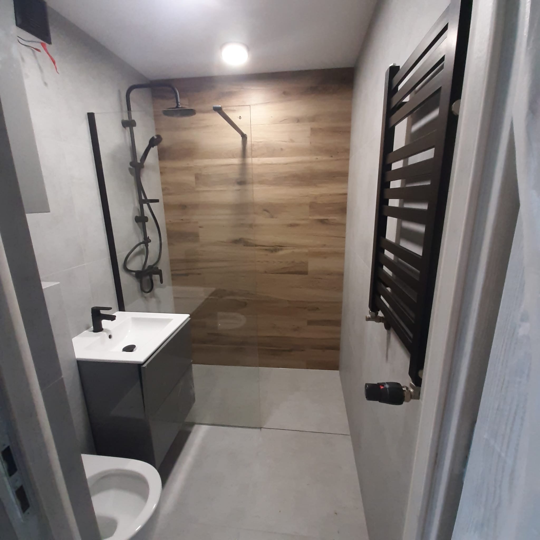 Nowoczesna łazienka z kabiną prysznicową typu walk-in, umywalką z szafką w kolorze grafitowym, grzejnikiem drabinkowym w czarnym macie oraz płytkami imitującymi drewno na jednej ze ścian.