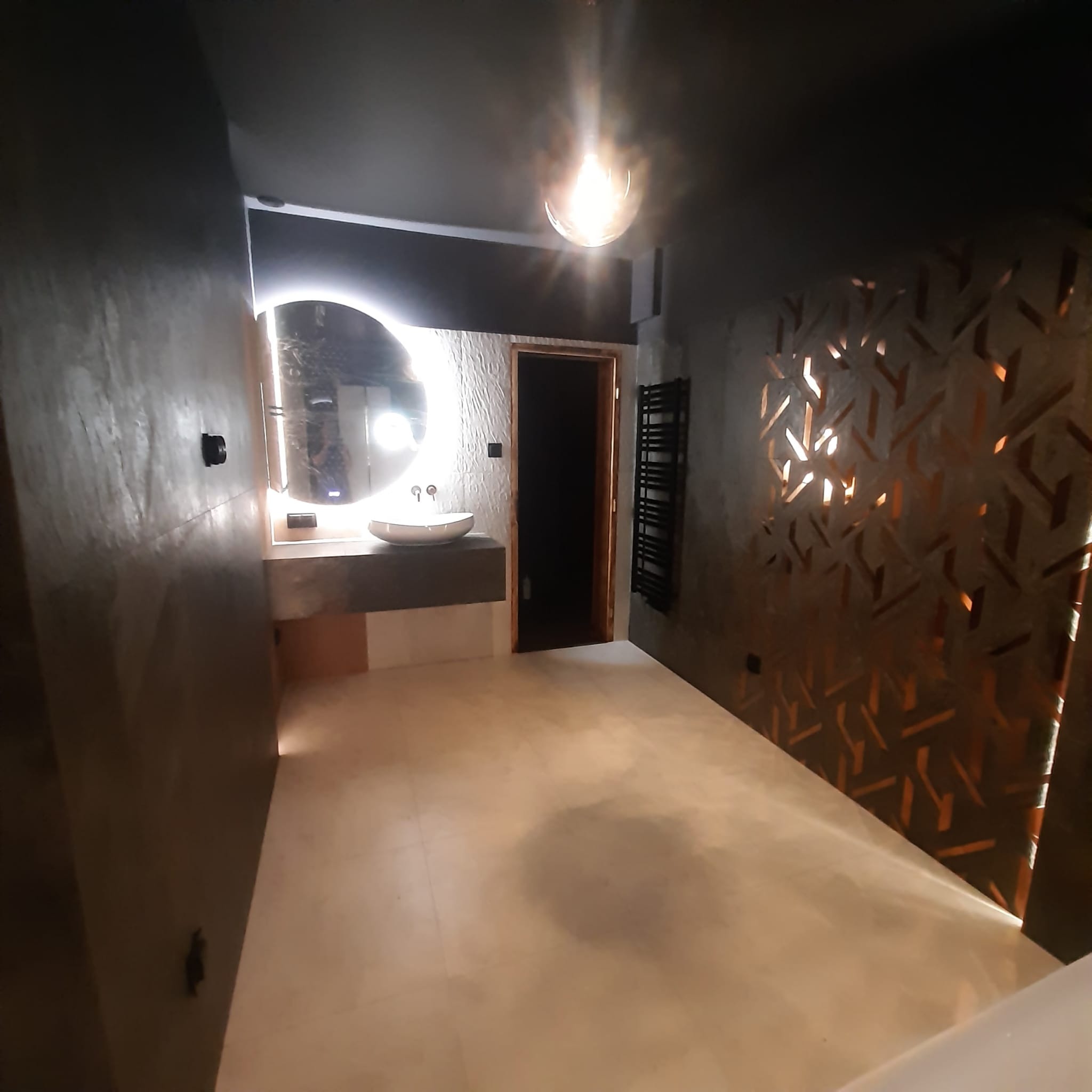 Nowoczesna łazienka z podświetlanym lustrem, umywalką nablatową na betonowym blacie, dekoracyjną ścianą z ażurowym wzorem i grzejnikiem drabinkowym w ciemnej tonacji.