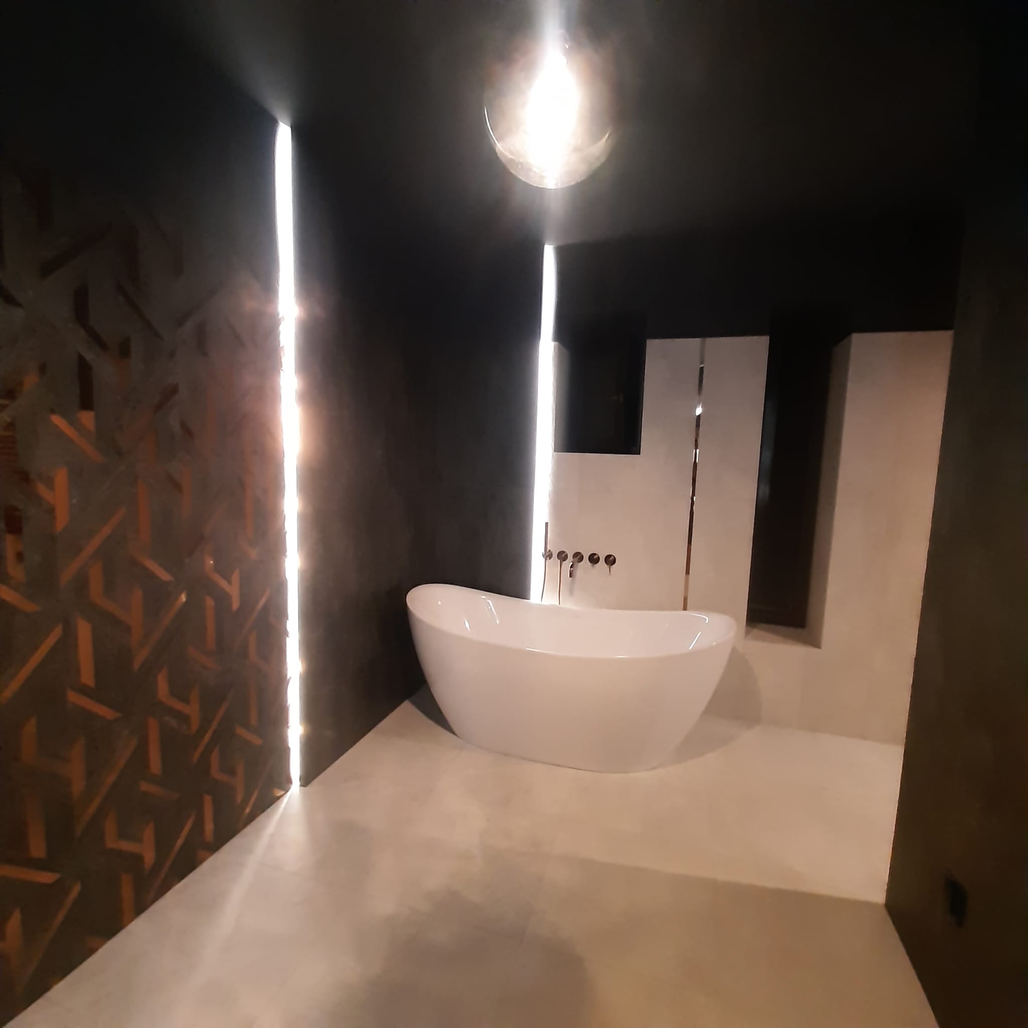 Nowoczesna łazienka z wolnostojącą białą wanną, ciemną ścianą z geometrycznym wzorem i pionowymi paskami świetlnymi.