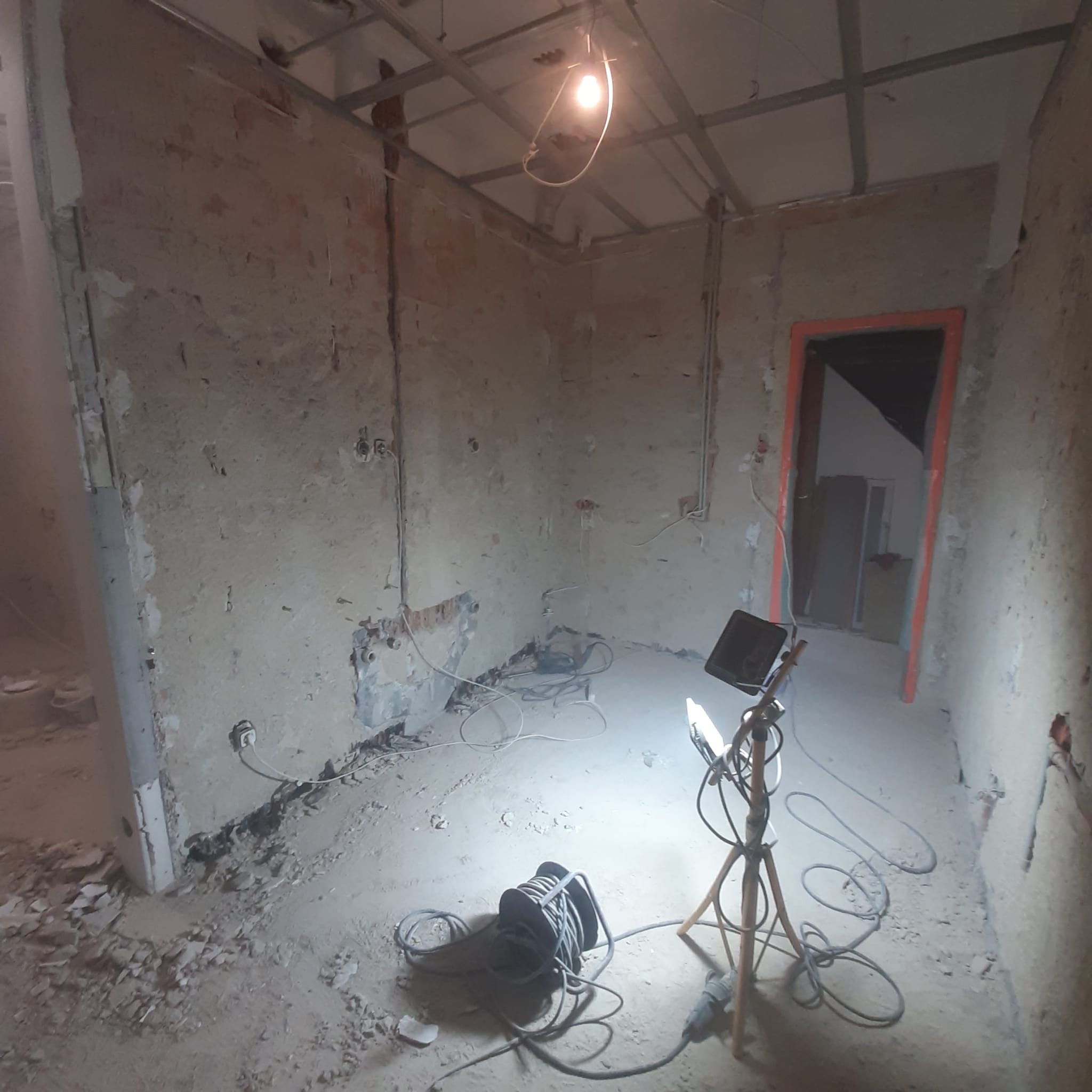 Wnętrze w stanie surowym podczas prac remontowych, widoczne ściany z odsłoniętą cegłą i instalacjami elektrycznymi, prowizoryczne oświetlenie budowlane oraz rozciągnięte przewody elektryczne...