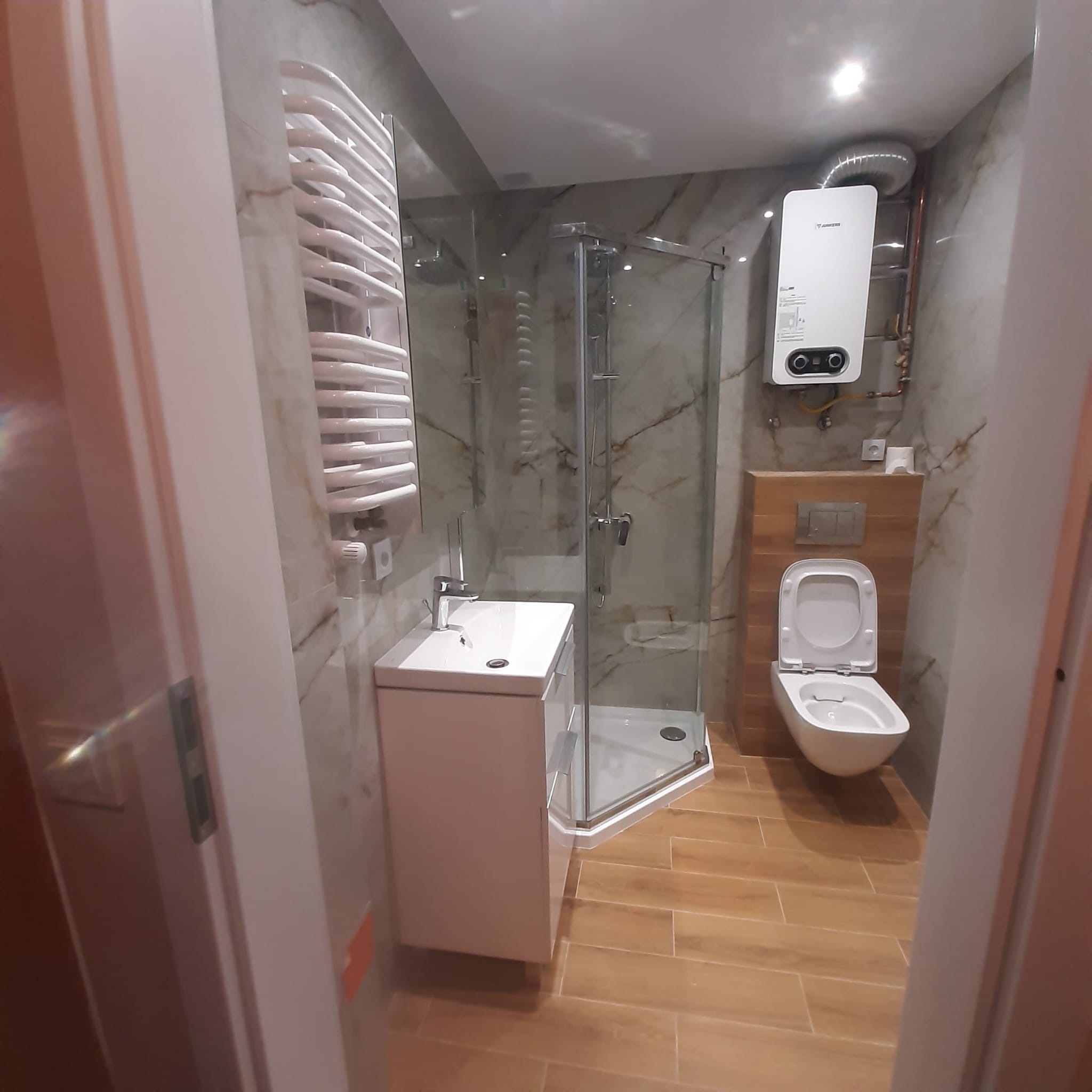 Nowoczesna łazienka po remoncie z kabiną prysznicową, umywalką z szafką, podwieszaną toaletą, grzejnikiem drabinkowym i piecem gazowym, wykończona płytkami imitującymi marmur i drewno.