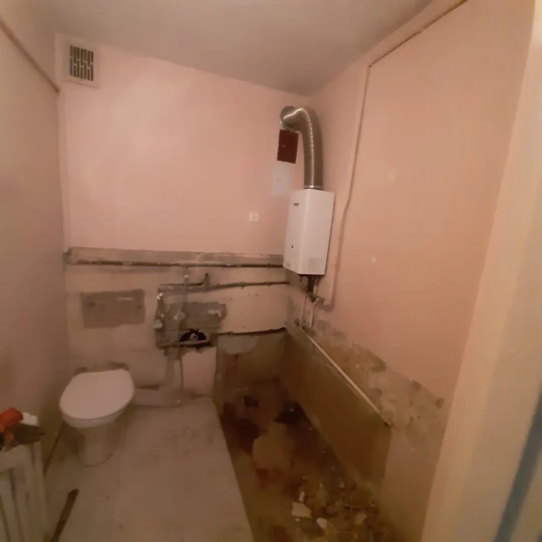 Demontaż płytek w łazience, widoczny fragment toalety, ściany w różowym kolorze, odsłonięte rury i instalacje, przygotowanie do dalszych prac remontowych.