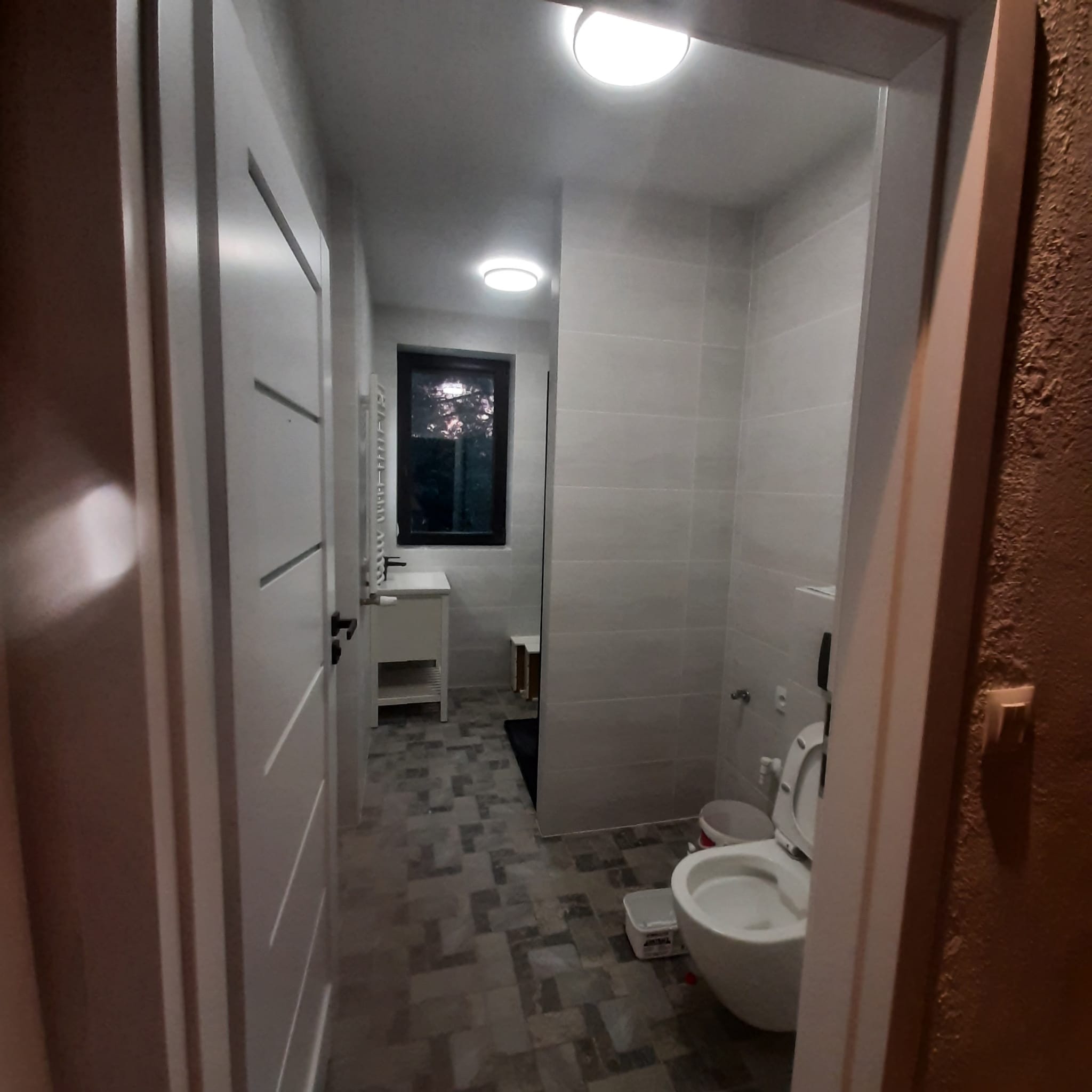 Wykończona łazienka z szarymi płytkami, widok przez otwarte drzwi z nowoczesną białą ościeżnicą, na podłodze wzorzyste płytki imitujące kamień, w tle okno.