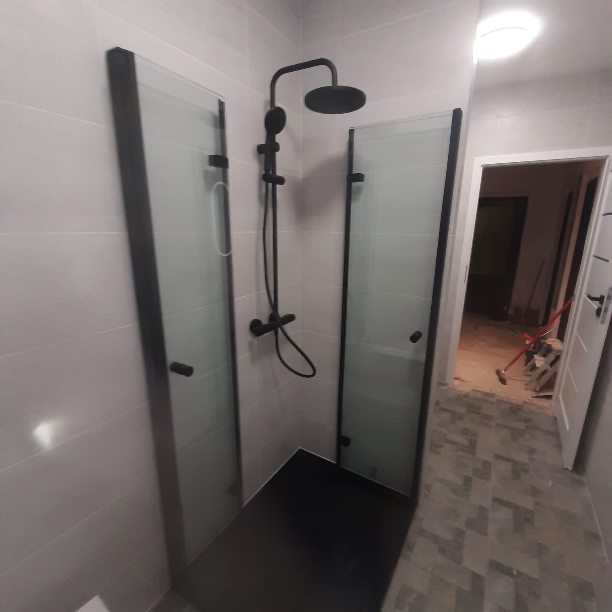 Kabina prysznicowa z czarną armaturą i matowymi szybami, widoczna w rogu łazienki z szarą glazurą. W tle widoczne prace wykończeniowe w pomieszczeniu obok.