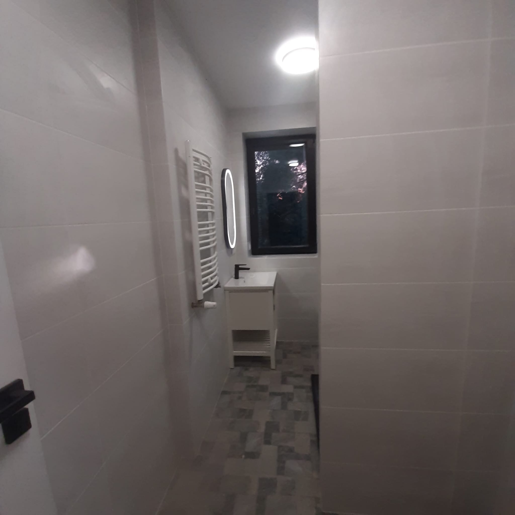 Nowoczesna, wąska łazienka z białymi, podłużnymi płytkami na ścianach, grzejnikiem drabinkowym, okrągłym lustrem z podświetleniem LED, białą szafką z umywalką i ciemną ramą okna z widokiem...