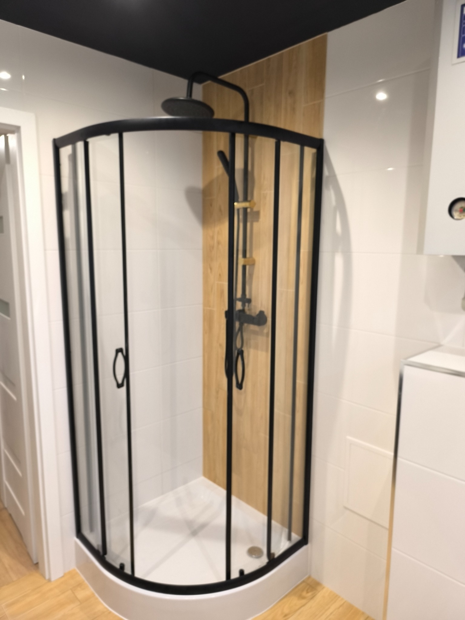 Narożna kabina prysznicowa z czarnymi profilami i armaturą w odcieniach czerni i złota, zamontowana w łazience z białymi płytkami i panelami imitującymi drewno.