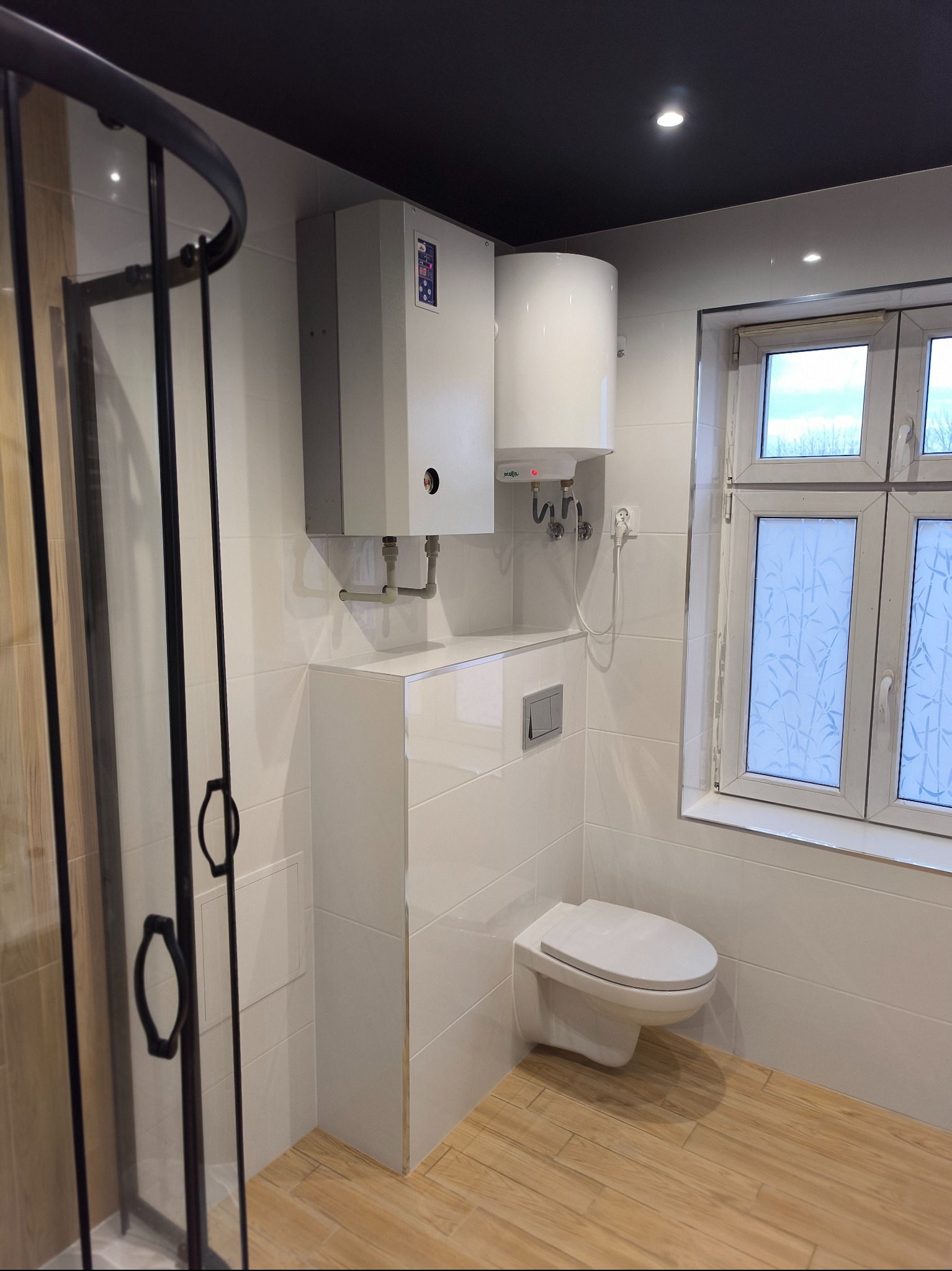 Nowoczesna łazienka z kabiną prysznicową z czarnymi profilami, podwieszaną toaletą, białymi płytkami i panelami podłogowymi imitującymi drewno; widoczny bojler i piec.