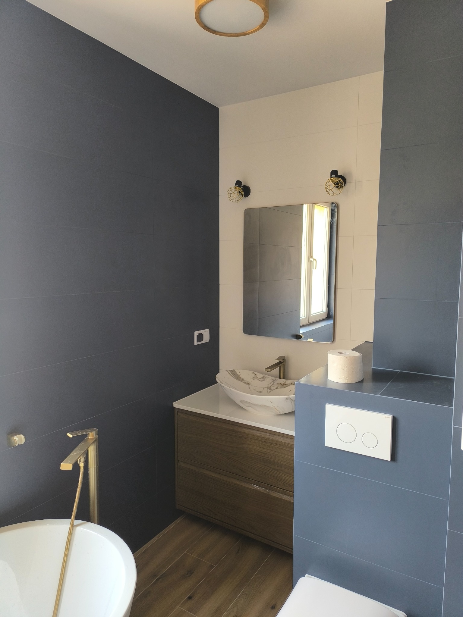 Nowoczesna łazienka z szarymi płytkami ściennymi, białą umywalką marmurkową na drewnianej szafce, złotą armaturą i lustrem z zaokrąglonymi rogami.