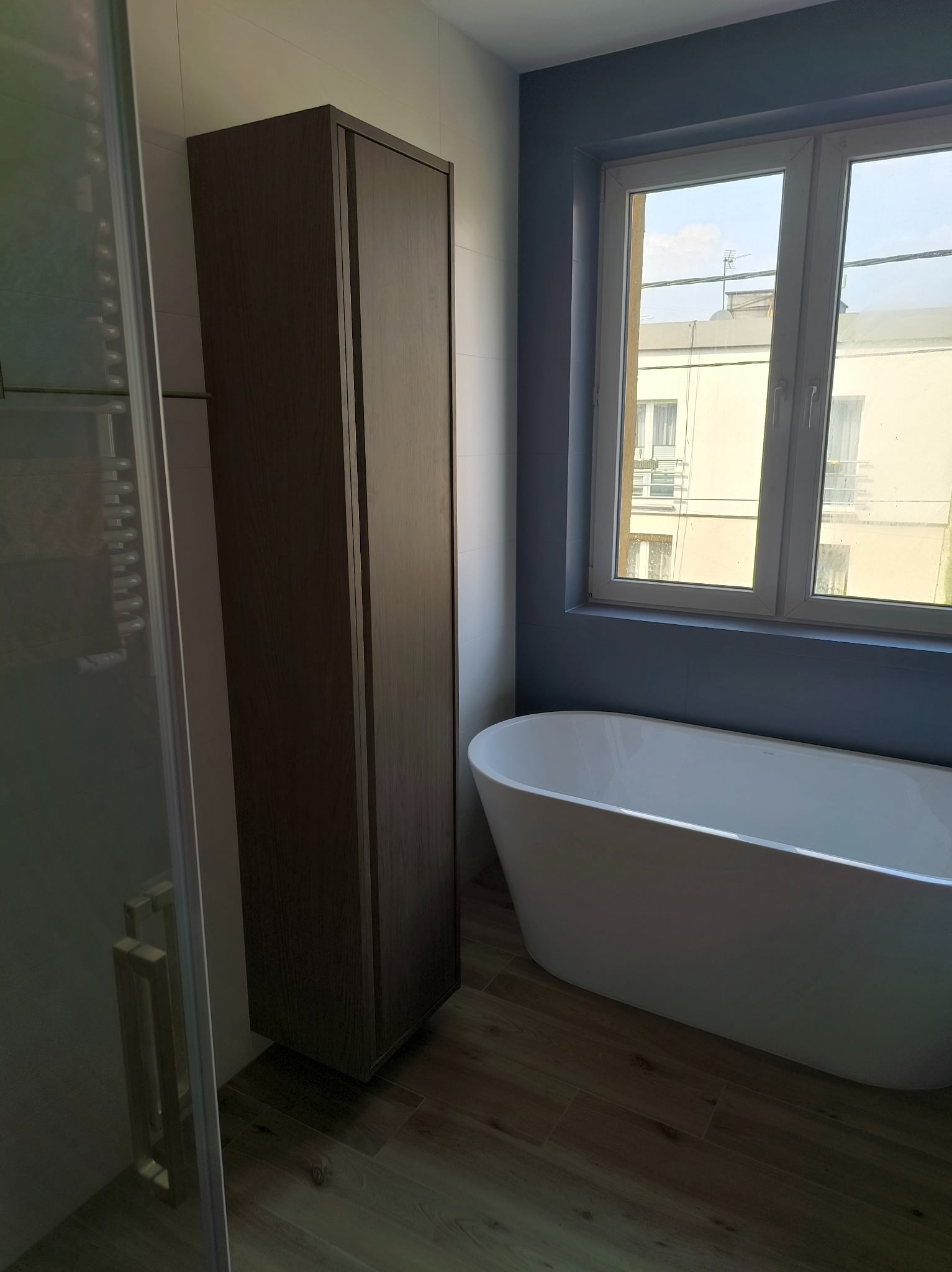 Nowoczesna łazienka z wolnostojącą białą wanną, wysoką szafką w kolorze ciemnego drewna, kabiną prysznicową ze szklanymi drzwiami i oknem z widokiem na budynek mieszkalny.