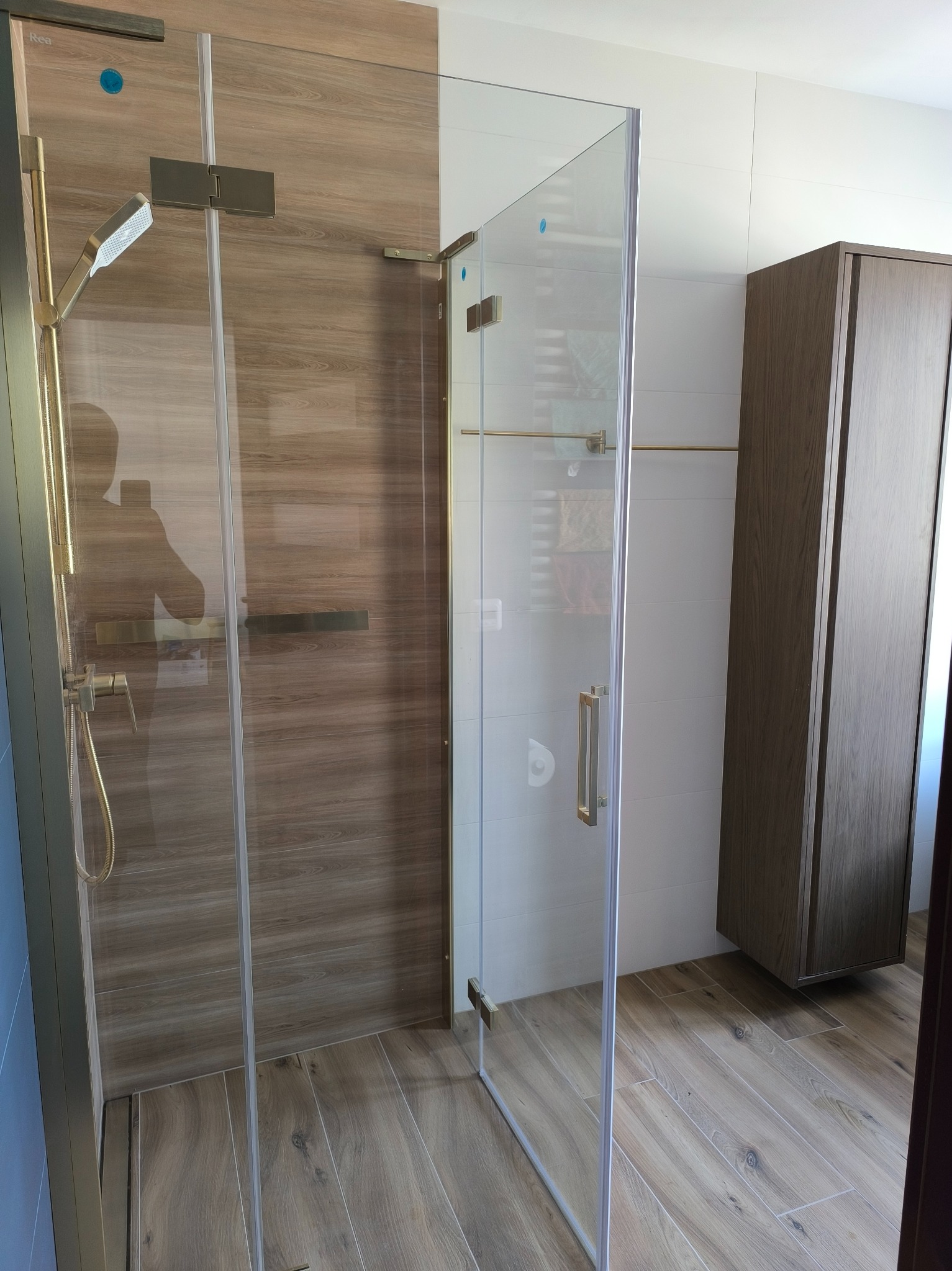 Nowoczesna kabina prysznicowa z elementami w kolorze złotym, drewnopodobne płytki na ścianach i podłodze, minimalistyczna szafka łazienkowa w tle.
