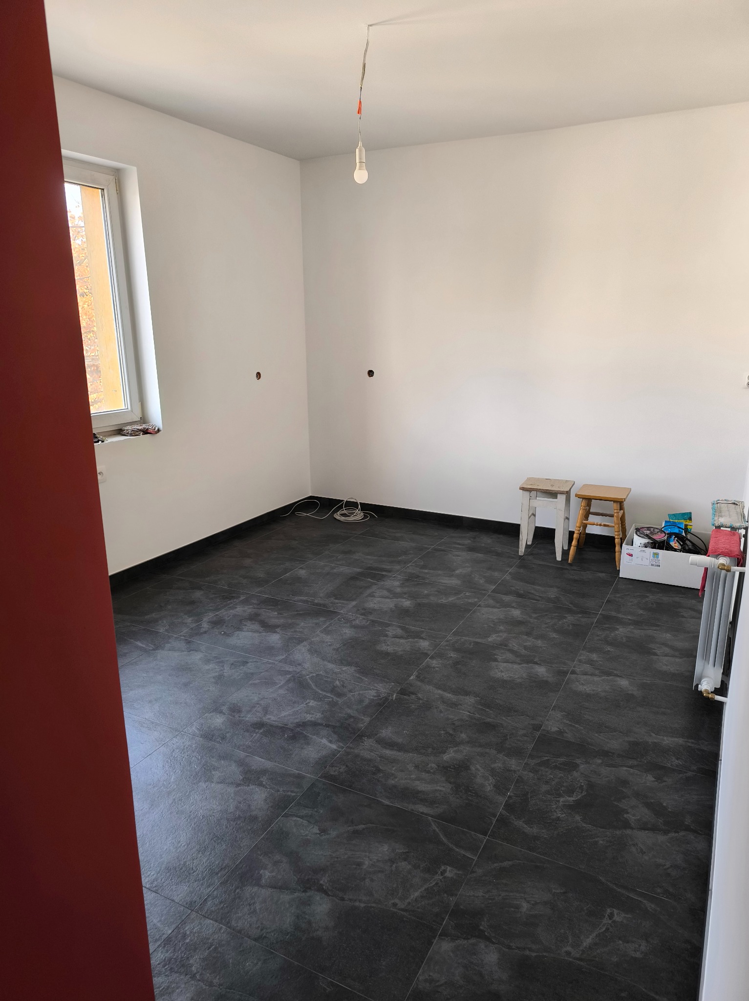 Pusty pokój po remoncie z ciemnymi płytkami podłogowymi, białymi ścianami i wiszącą żarówką. Dwa drewniane stołki i karton z akcesoriami w rogu pokoju.