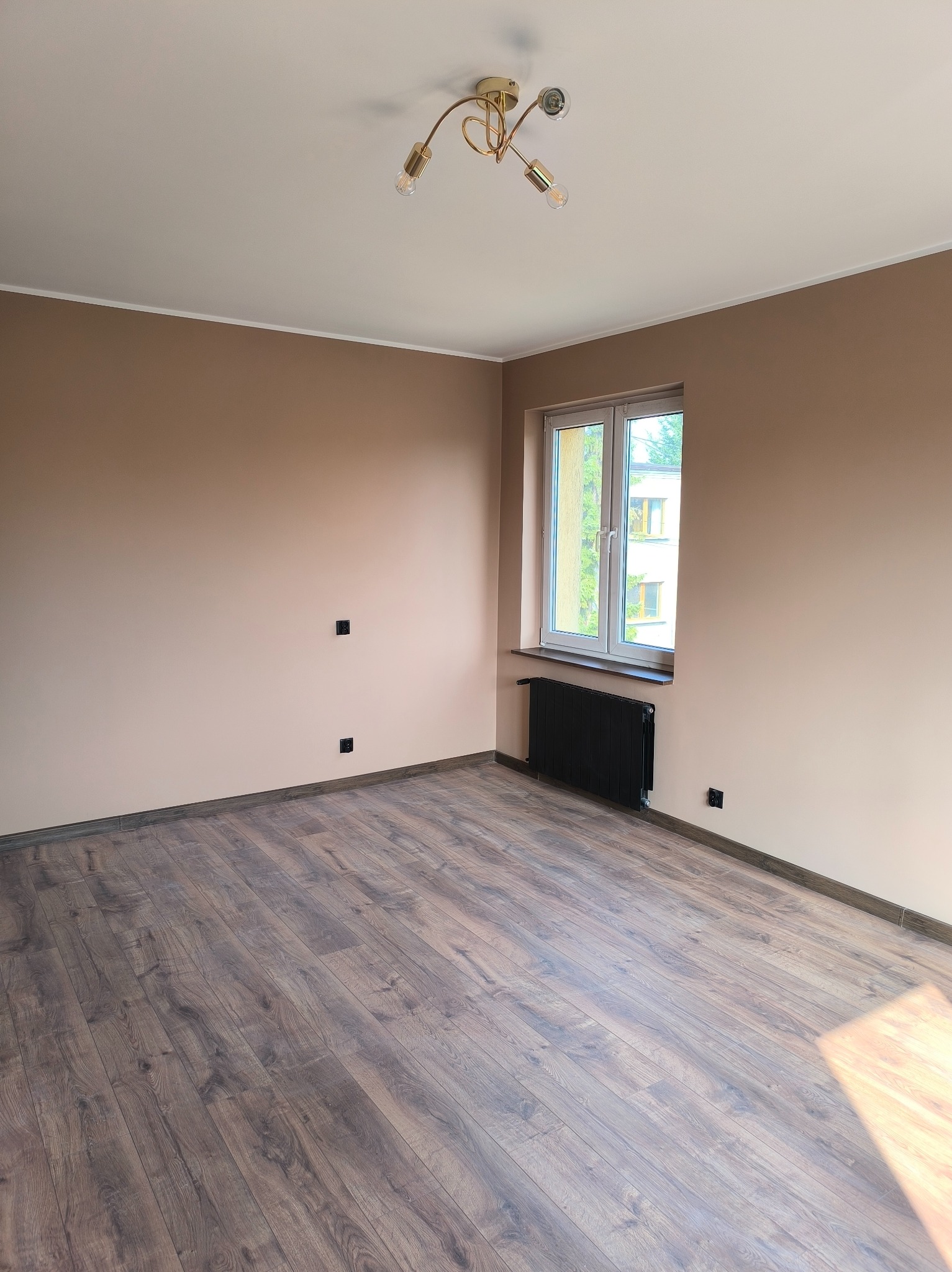 Pusty pokój po odświeżeniu: ściany pomalowane na ciepły beż, ciemne panele podłogowe, białe okno z czarnym grzejnikiem pod spodem, złoty żyrandol na suficie, widoczne gniazdka elektryczne.