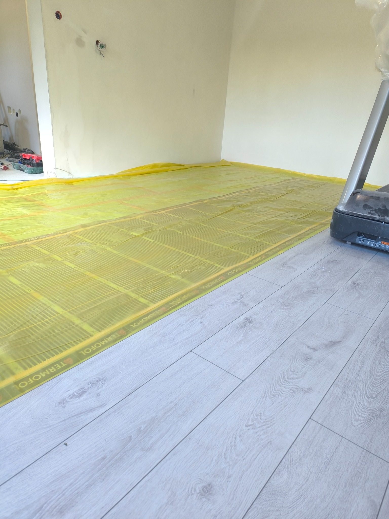Montaż ogrzewania podłogowego: żółta folia grzewcza Termofol rozłożona na podłożu, częściowo zakryta panelami podłogowymi w jasnym odcieniu drewna.