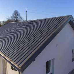 Profesjonalne.usługi.dekarskie - Dom jednorodzinny z nowym, ciemnoszarym dachem z blachy trapezowej, widok z góry, błękitne niebo w tle.