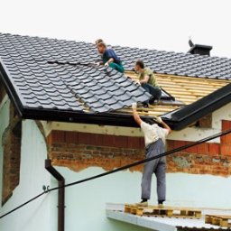 Profesjonalne.usługi.dekarskie - Trzech dekarzy układa ciemnoszarą dachówkę na drewnianej konstrukcji dachu domu jednorodzinnego z widoczną elewacją w trakcie remontu.