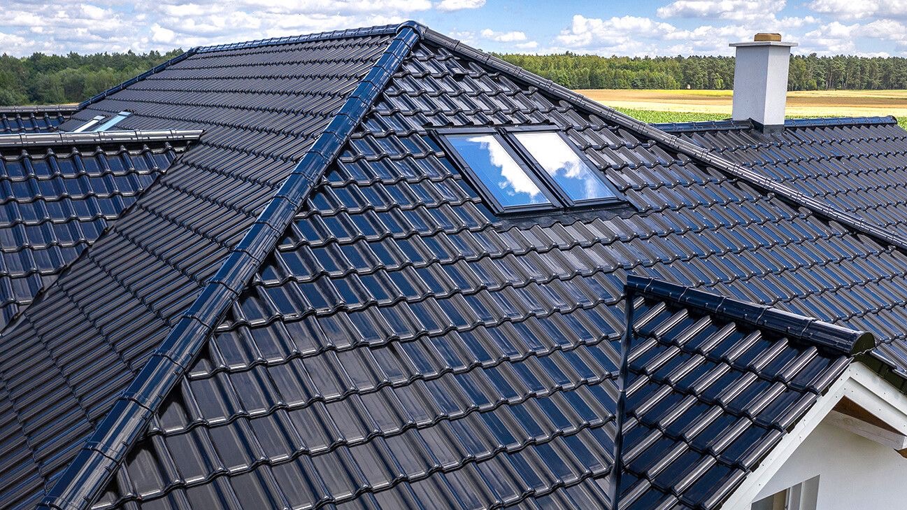 Błyszczący, czarny dach z dachówki ceramicznej z dwoma oknami dachowymi, widok na komin i zielone pola w tle.