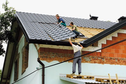 Trzech dekarzy układa ciemnoszarą dachówkę na drewnianej konstrukcji dachu domu jednorodzinnego z widoczną elewacją w trakcie remontu.