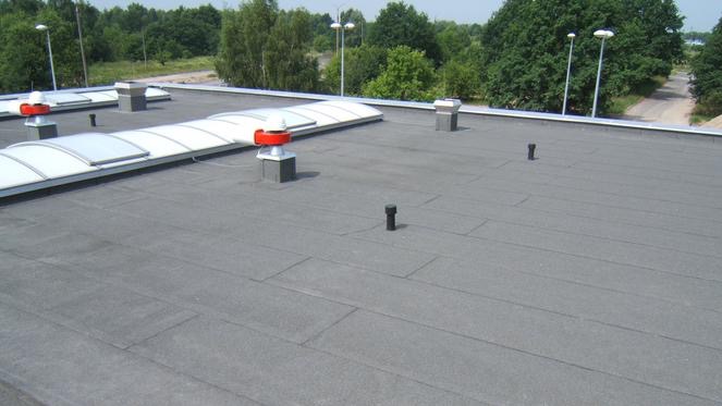 Płaski dach pokryty papą termozgrzewalną z kominkami wentylacyjnymi, świetlikami dachowymi i uszczelnieniami.