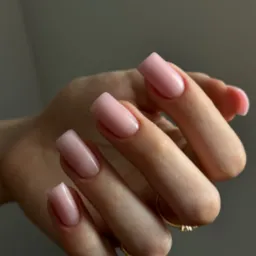 Dłoń z delikatnym, różowym manicurem hybrydowym o kwadratowym kształcie paznokci, złoty pierścionek na palcu serdecznym, neutralne tło.