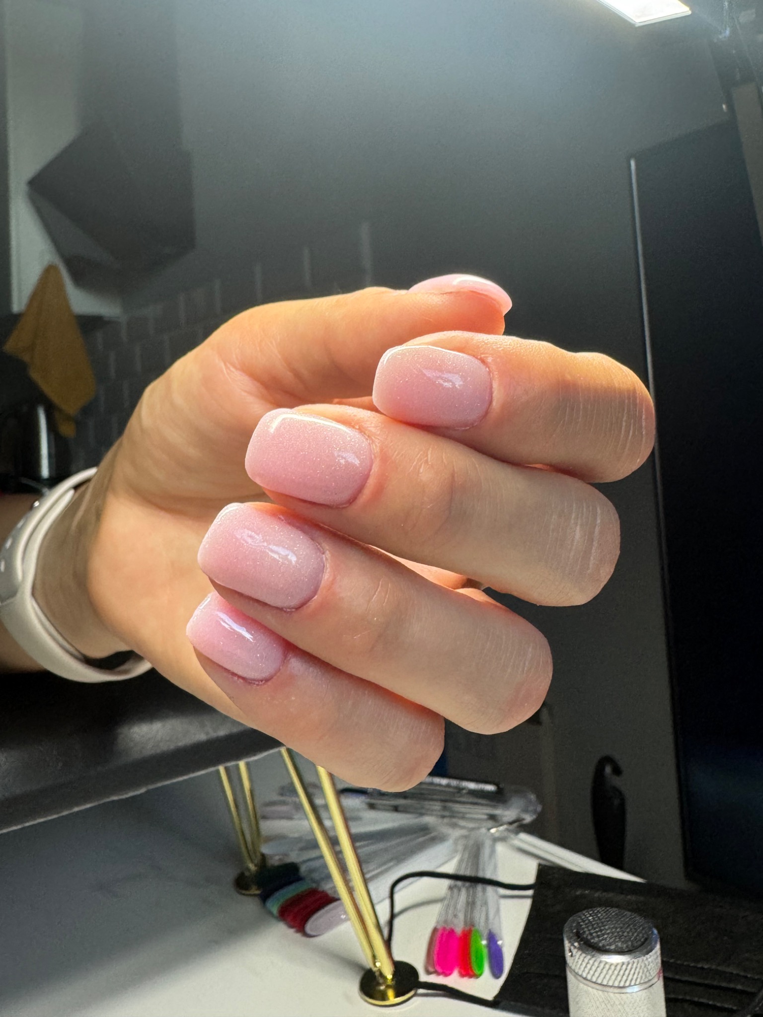 Dłoń z delikatnym, różowym manicure hybrydowym o kwadratowych paznokciach, widoczne próbki kolorów na stole.