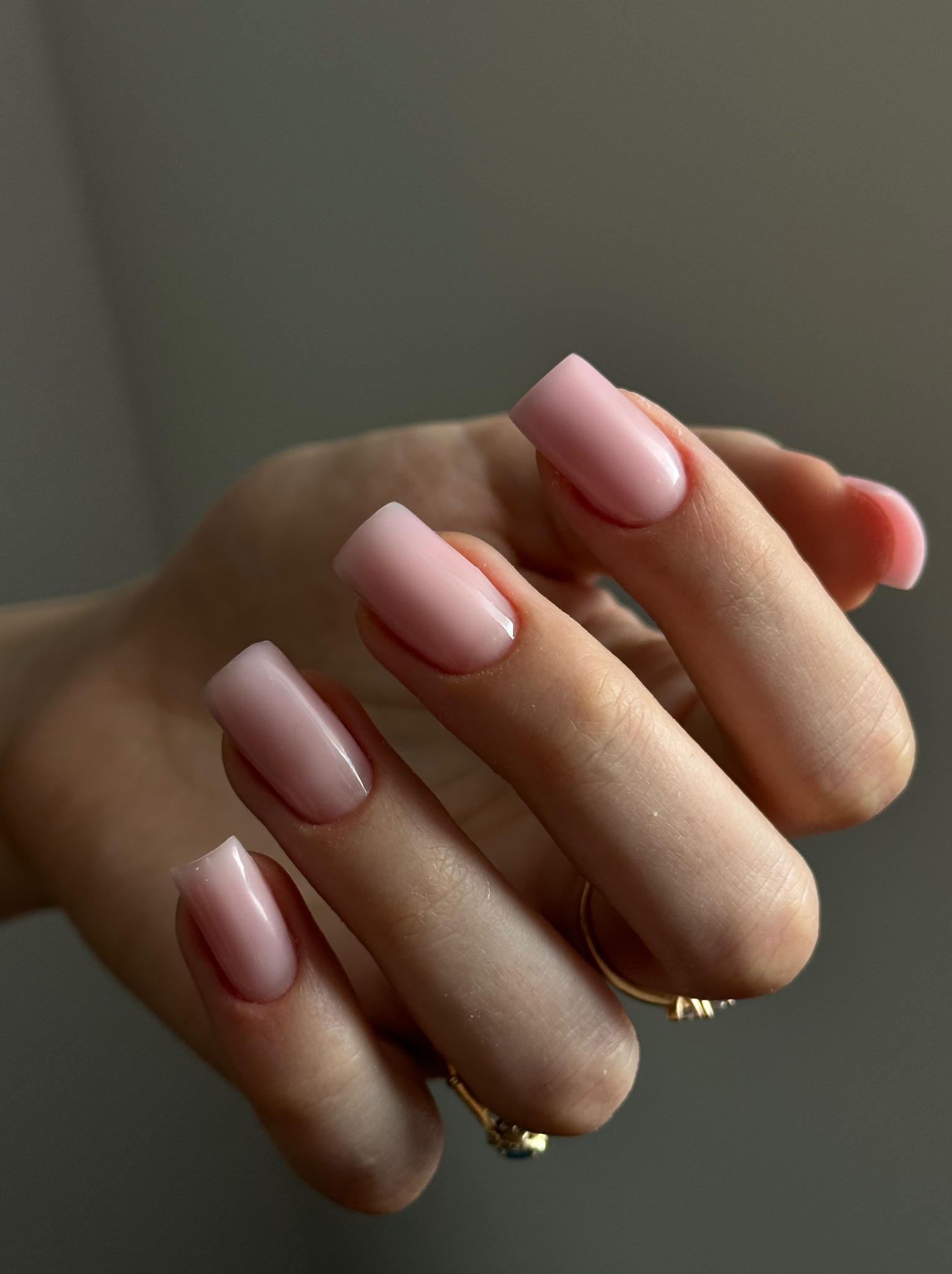 Dłoń z delikatnym, różowym manicurem hybrydowym o kwadratowym kształcie paznokci, złoty pierścionek na palcu serdecznym, neutralne tło.
