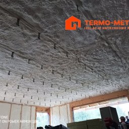 Termo-metr Tomasz Kudła - Poddasze domu jednorodzinnego po natrysku szarej pianki izolacyjnej, widoczne metalowe kołki mocujące i fragment logo firmy 'TERMO-METR' w prawym górnym rogu.