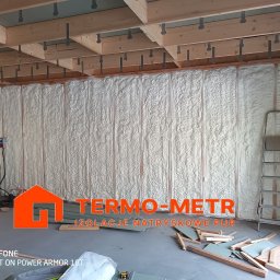 Termo-metr Tomasz Kudła - Ściana z drewnianą konstrukcją wypełniona białą pianką izolacyjną PUR, widoczna drabina oparta o ścianę, resztki materiałów budowlanych na podłodze, pracownik w oknie w oddali.