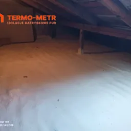 Poddasze ocieplone białą pianką PUR, widoczne drewniane elementy konstrukcji dachu oraz logo firmy 'TERMO-METR' w lewym górnym rogu.