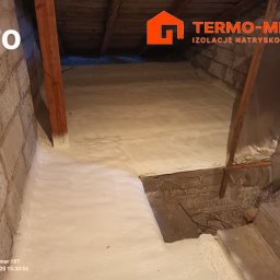 Termo-metr Tomasz Kudła - Poddasze po natrysku białej piany izolacyjnej, widoczne drewniane elementy konstrukcyjne i fragment ściany z cegły, z logo firmy TerMo-Metr w prawym górnym rogu i napisem 'Po' w lewym górnym rogu.