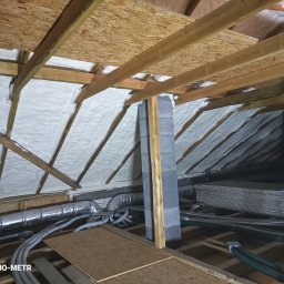 Termo-metr Tomasz Kudła - Poddasze z drewnianą konstrukcją i szarą pianką izolacyjną, widoczne przewody wentylacyjne i komin z bloczków. Ujęcie z dołu, z widocznym logo firmy w rogu.