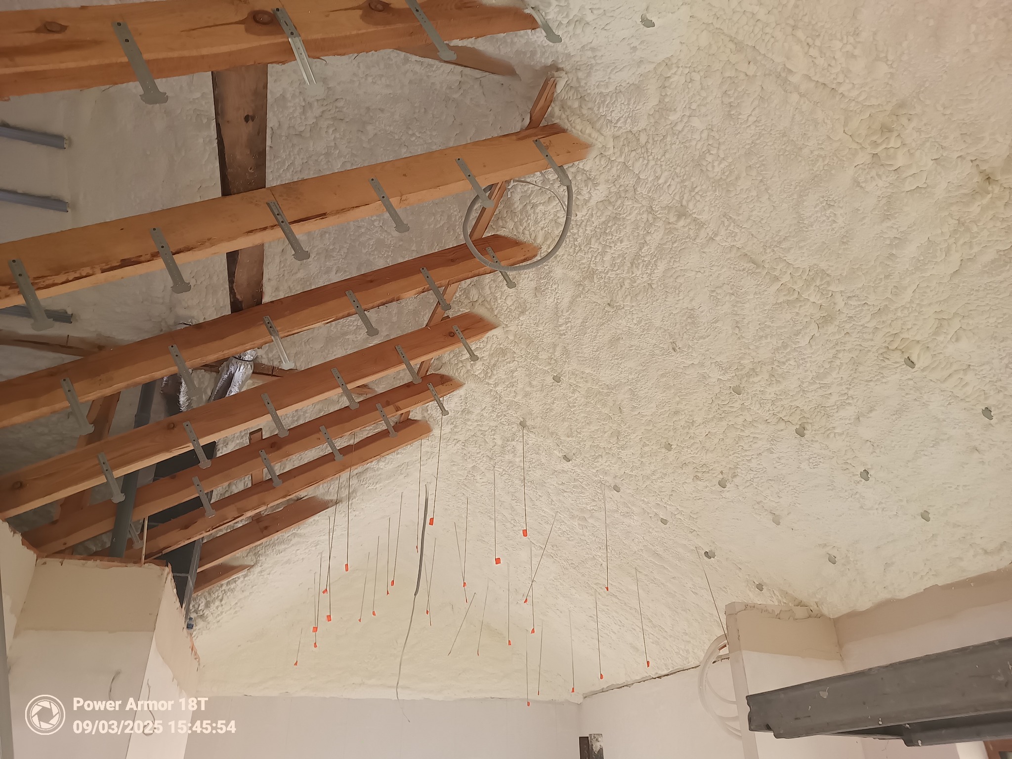 Poddasze z drewnianą konstrukcją po ociepleniu pianką PUR, widoczne wystające przewody z pomarańczowymi znacznikami. Biała, nierówna powierzchnia izolacji.