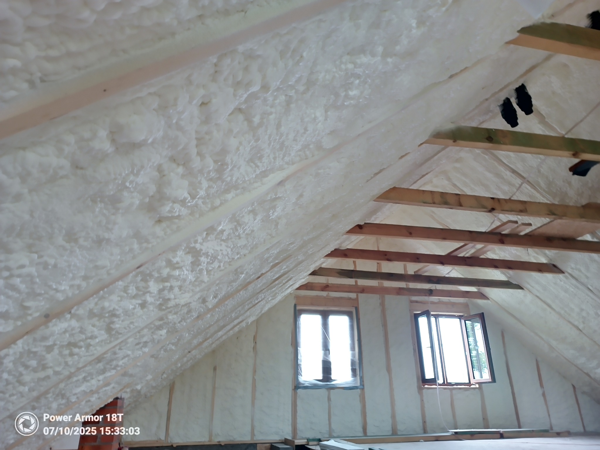 Poddasze po ociepleniu pianką PUR, widoczne drewniane elementy konstrukcyjne i okna. Izolacja pokrywa skosy dachu i ściany, tworząc jednolitą, jasną powierzchnię.