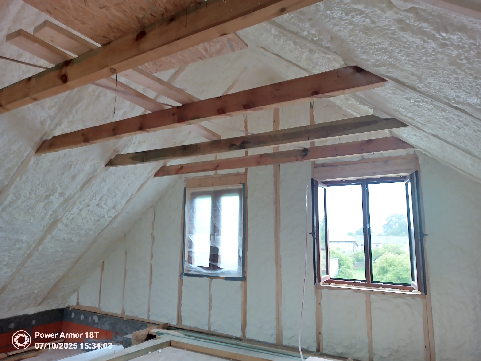 Poddasze po ociepleniu pianką PUR, widoczne drewniane belki stropowe i okna, ściany pokryte białą pianą izolacyjną o nieregularnej strukturze.