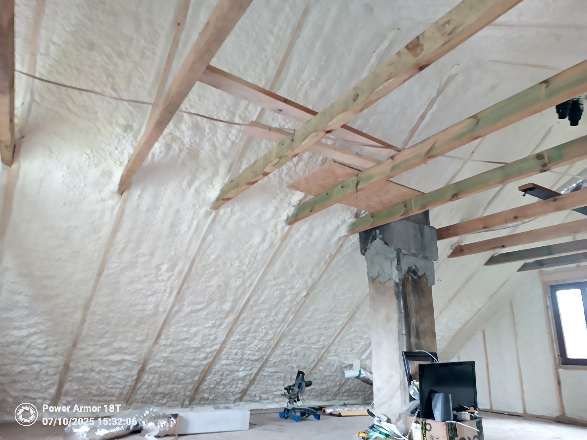 Poddasze po ociepleniu pianką, widoczne drewniane krokwie i komin. Na podłodze narzędzia i resztki materiałów. Oświetlenie naturalne z okna.