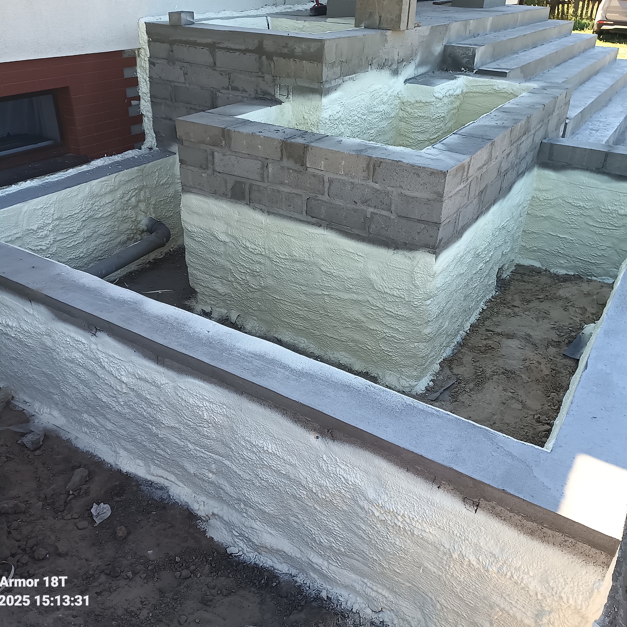 Fundamenty budynku w trakcie izolacji pianką PUR, widoczne bloczki betonowe i fragment rury. Prace izolacyjne w toku, nierównomierna struktura pianki.