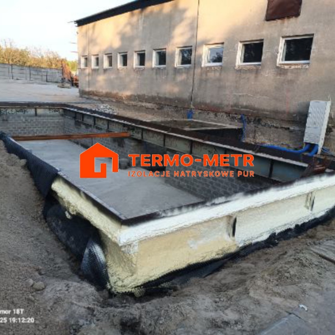 Izolacja fundamentu budynku przemysłowego pianką PUR, widoczna warstwa piany i folia ochronna. Ujęcie z ziemi, z widocznym fragmentem elewacji i okien.