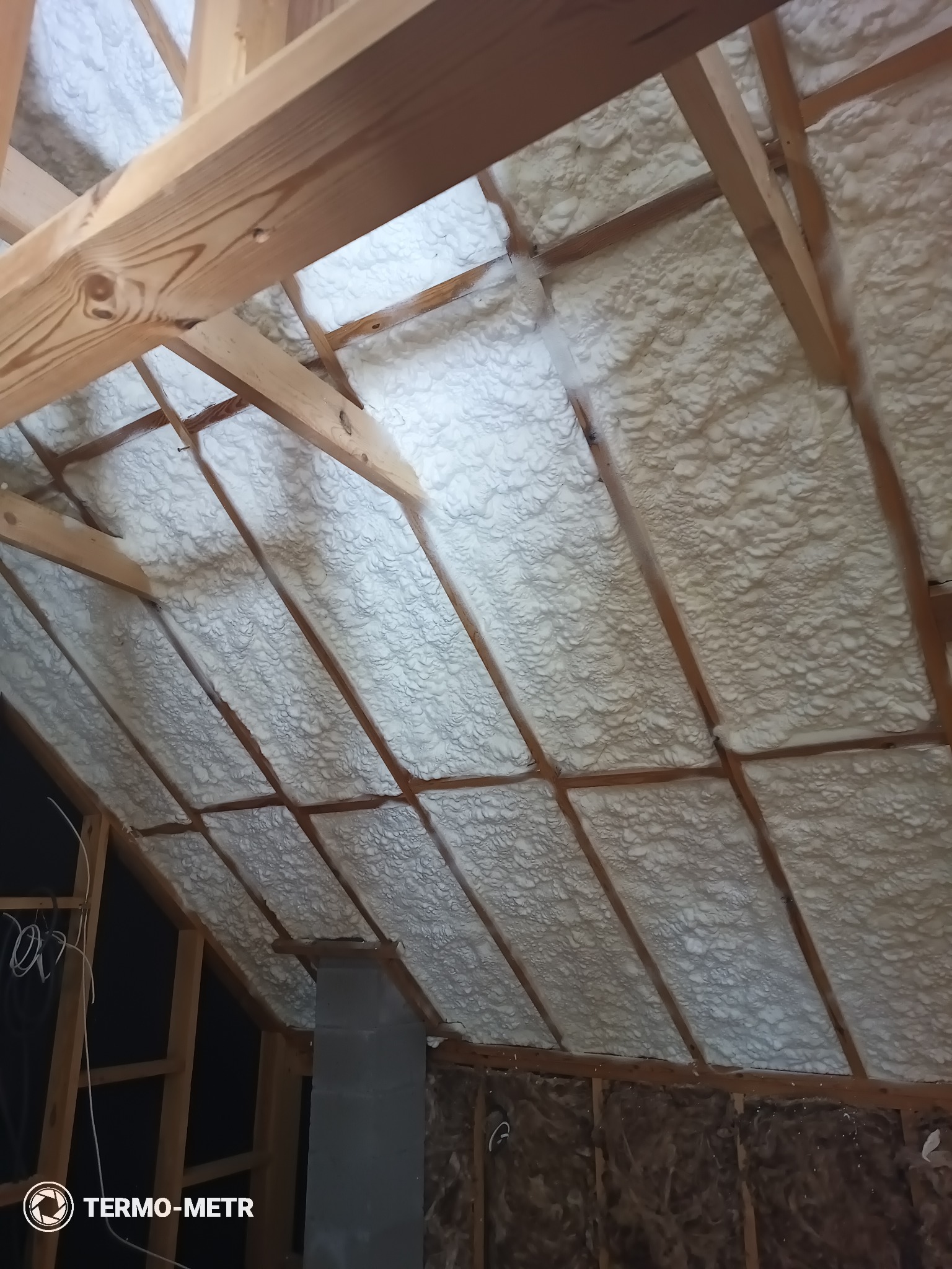 Poddasze z drewnianą konstrukcją pokryte białą pianką izolacyjną, widoczne przewody i fragment komina. Ukośny kadr prezentuje strukturę pianki i dokładność aplikacji.